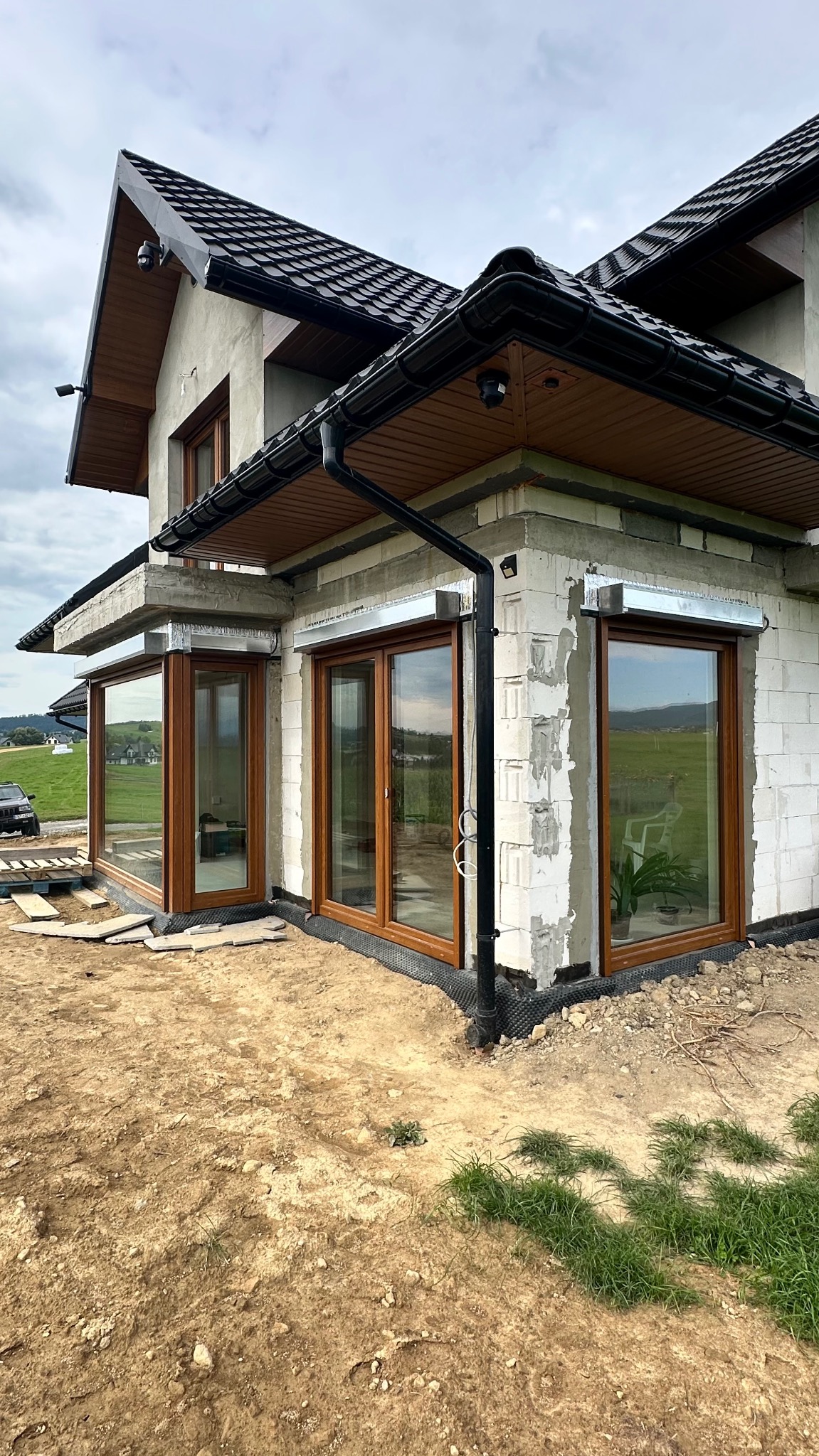 Surowy stan budynku z oknami PCV w kolorze drewna i przygotowanymi miejscami pod rolety zewnętrzne, widok z zewnątrz na tle krajobrazu.