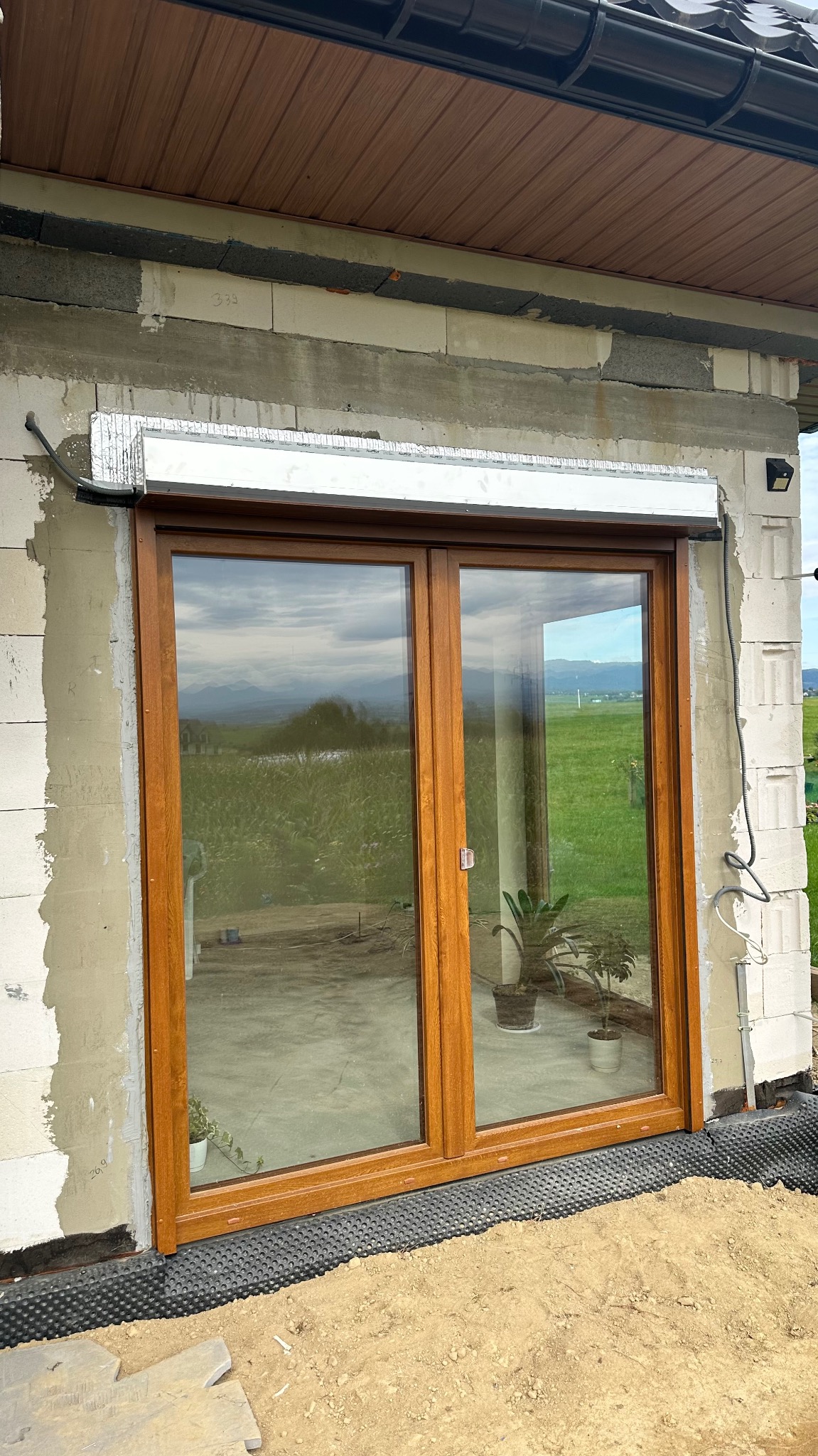 Montaż okna PCV z roletą zewnętrzną w surowym stanie budynku. Widoczne bloczki i ocieplenie. Za oknem krajobraz z górami.