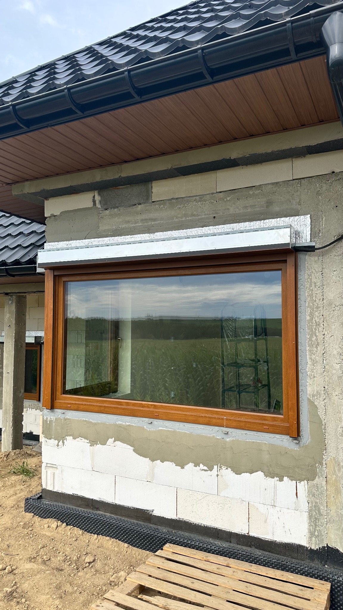 Brązowe okno PCV z zamontowaną roletą zewnętrzną na surowej ścianie budynku w trakcie budowy. Widoczne rusztowanie w odbiciu szyby.