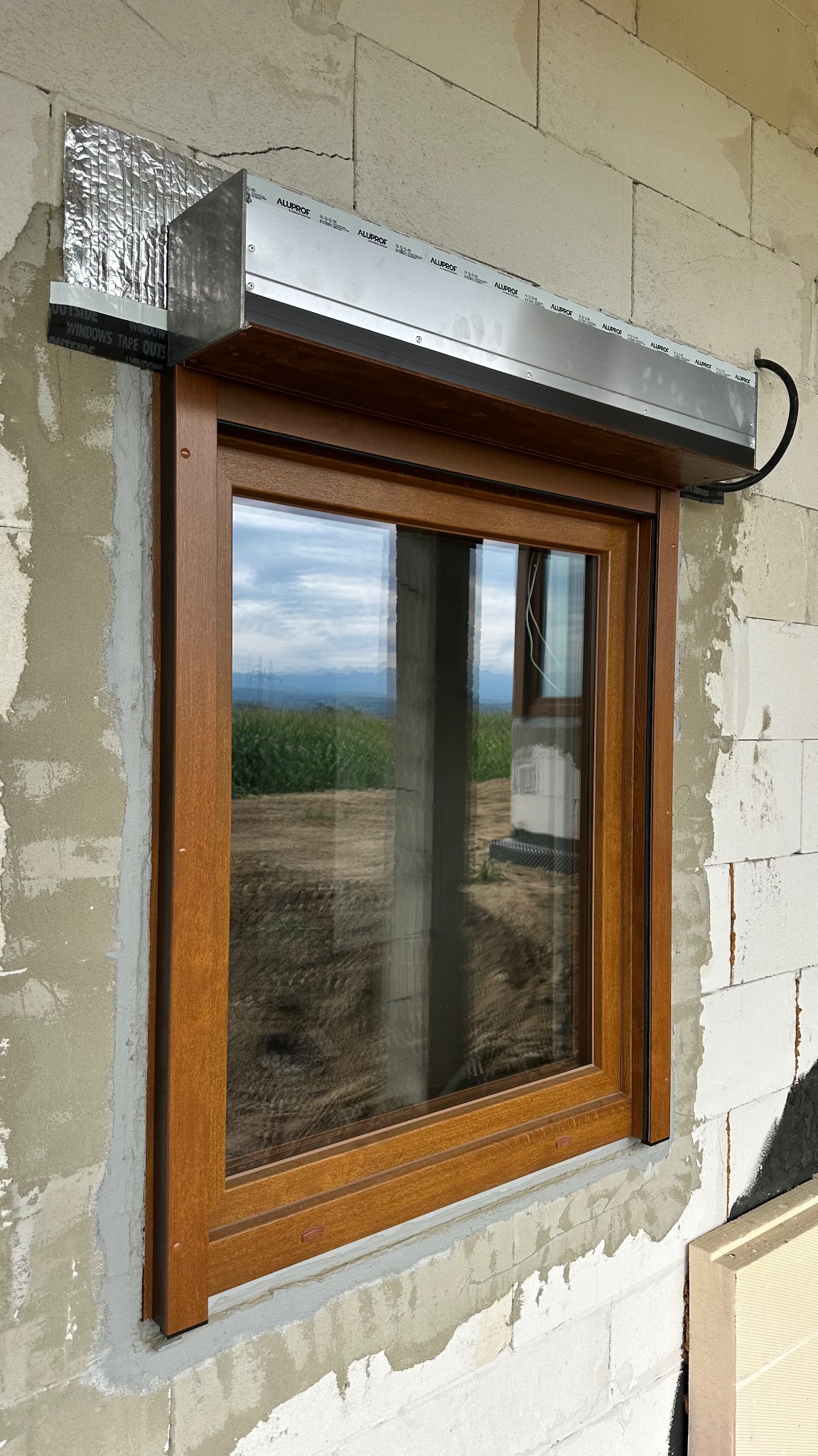 Drewnopodobne okno z roletą zewnętrzną w trakcie montażu, widoczne materiały izolacyjne i surowy mur. W szybie odbija się krajobraz.