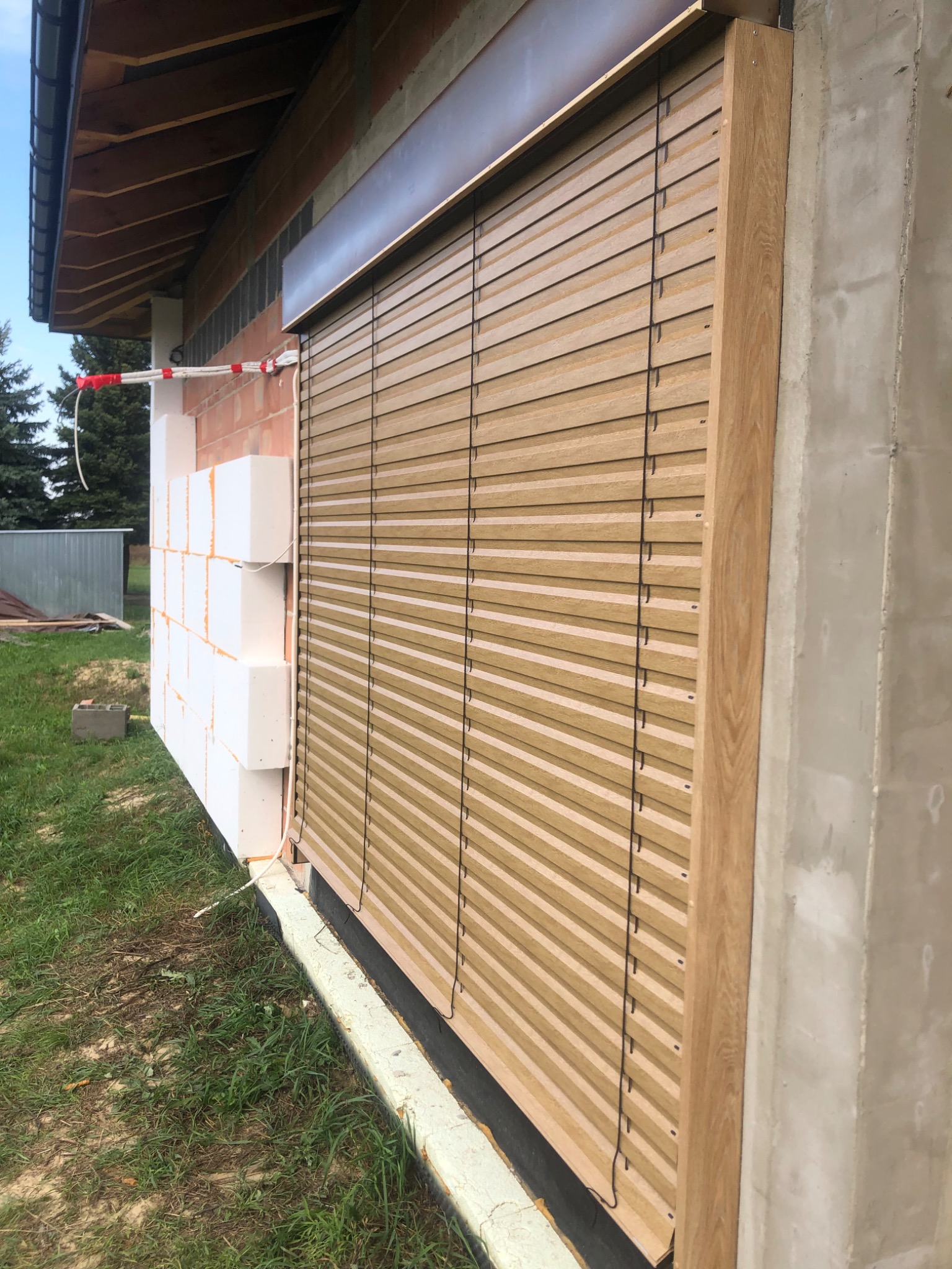 Zamknięte żaluzje fasadowe w kolorze drewna, zamontowane na budynku w trakcie budowy, z widocznym ociepleniem i fragmentem elewacji.