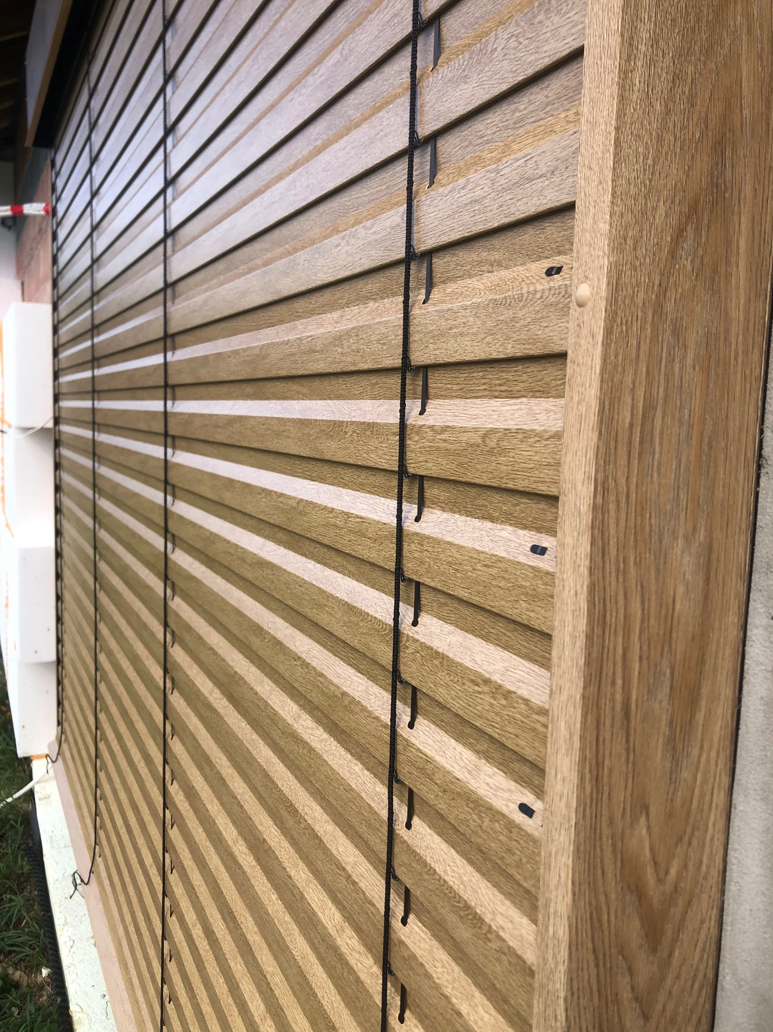 Zewnętrzne żaluzje drewniane w kolorze jasnego dębu, z czarnymi sznurkami, zamontowane na budynku z ociepleniem. Ujęcie pod kątem, ukazujące strukturę drewna.