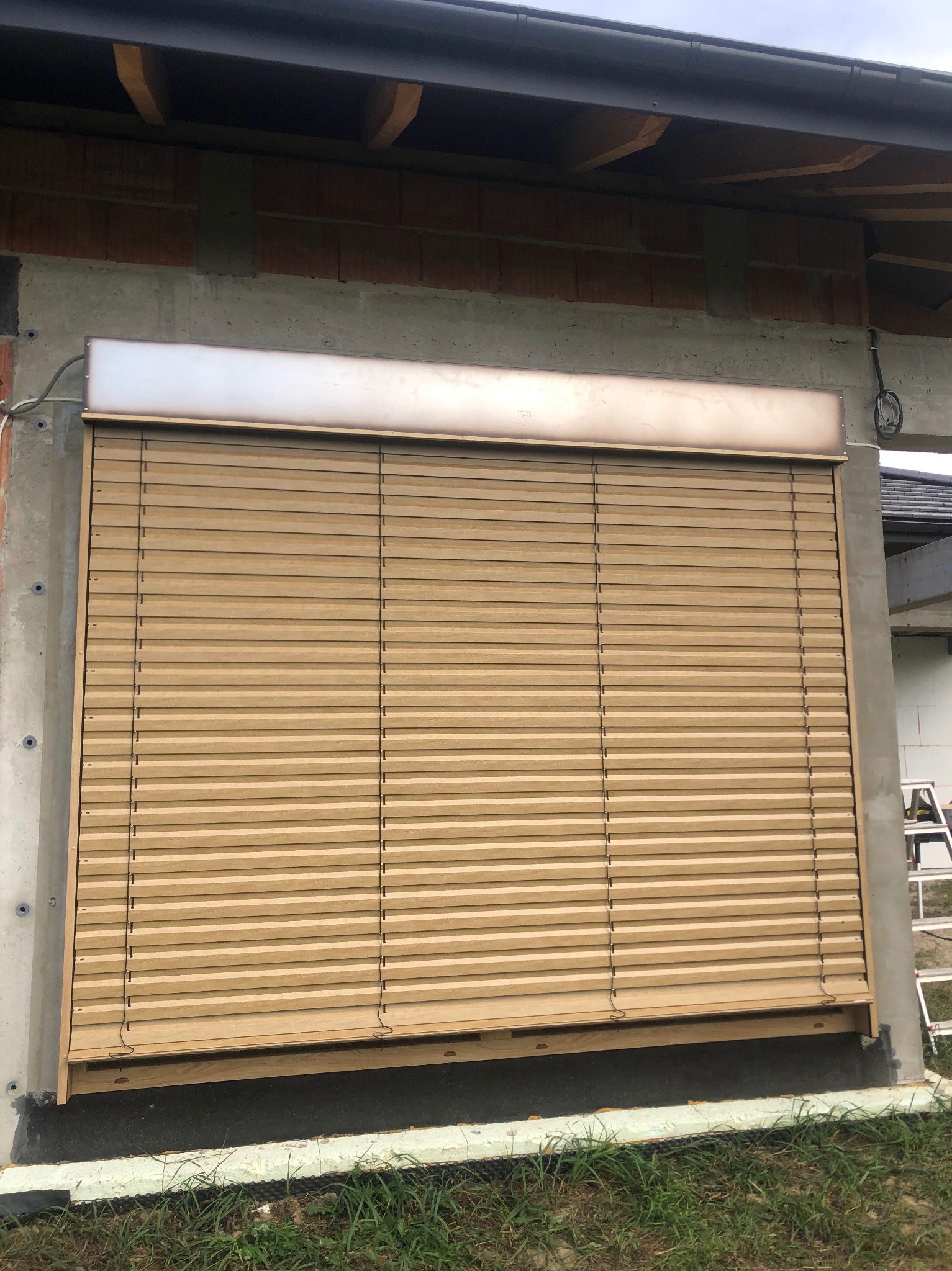 Zamknięta, beżowa roleta zewnętrzna na tle surowego muru, widoczna instalacja elektryczna. Detal: niedokończone wykończenie budynku w tle.