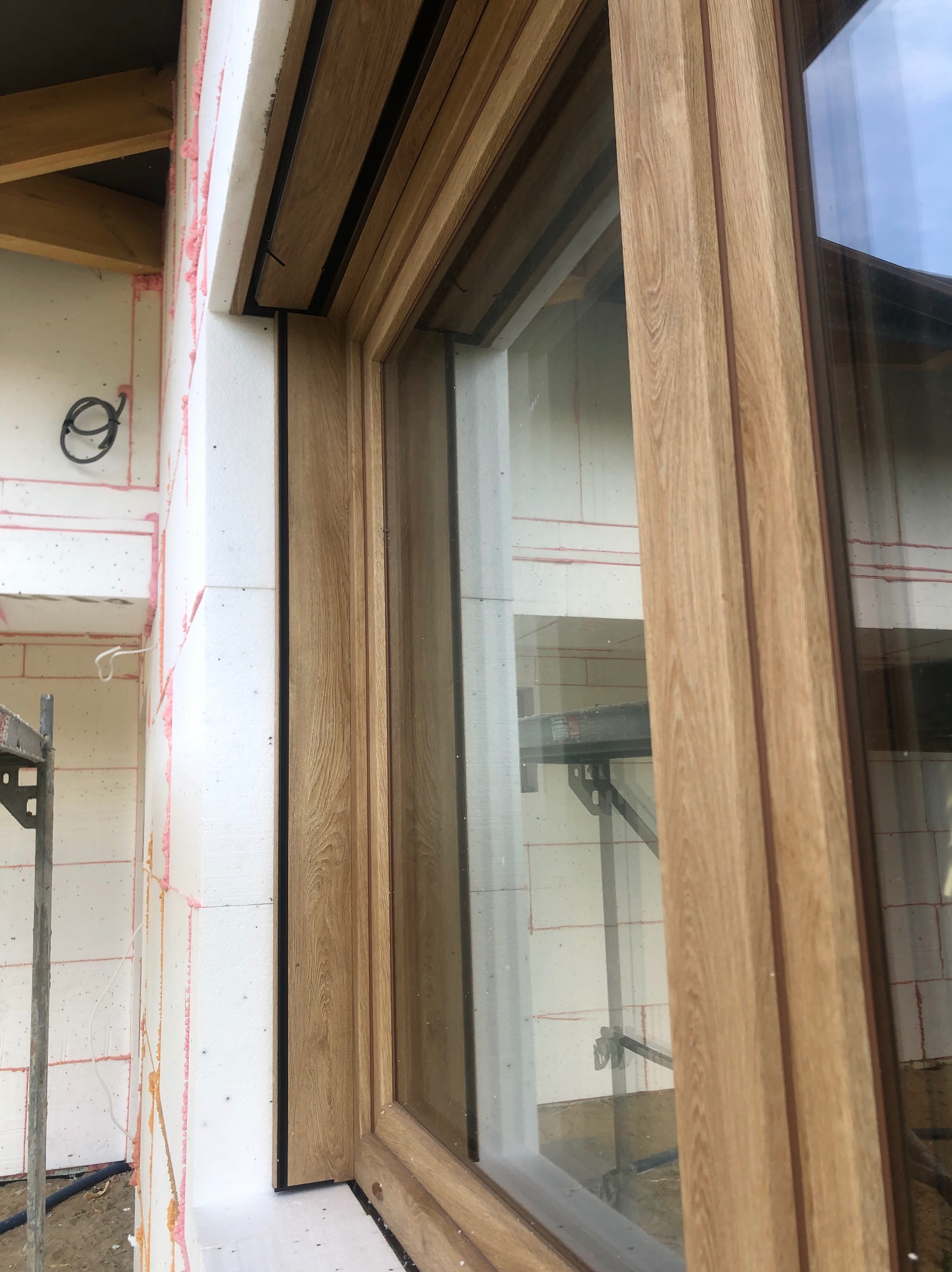 Drewnopodobne okno w trakcie montażu, widoczne ocieplenie budynku. Detal okna z bliska, na tle ściany z białego styropianu i fragment rusztowania.