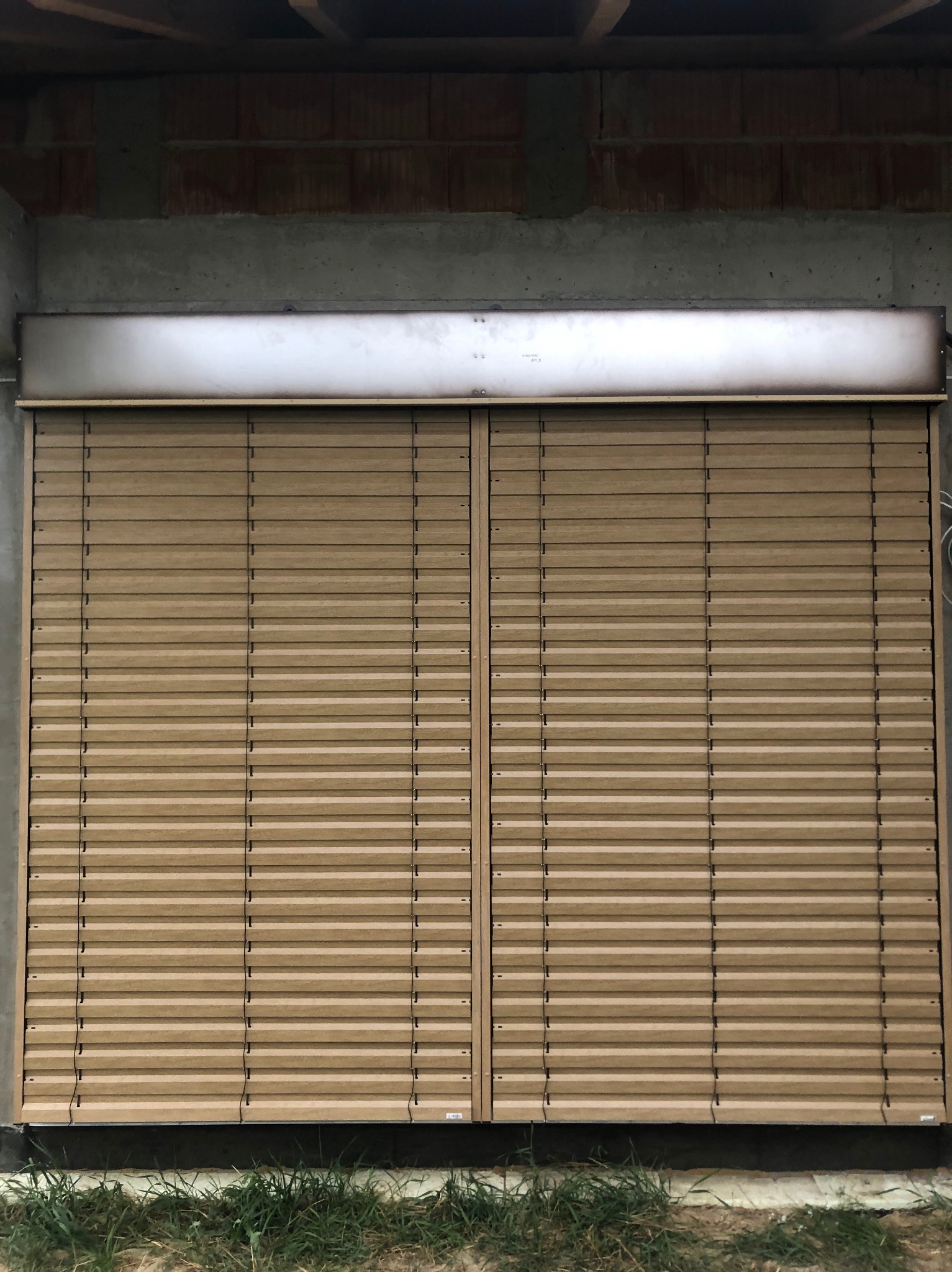 Zamknięte, beżowe żaluzje fasadowe na tle surowej ściany z betonu i cegieł. Widoczne elementy montażowe i fragment trawy u dołu zdjęcia.