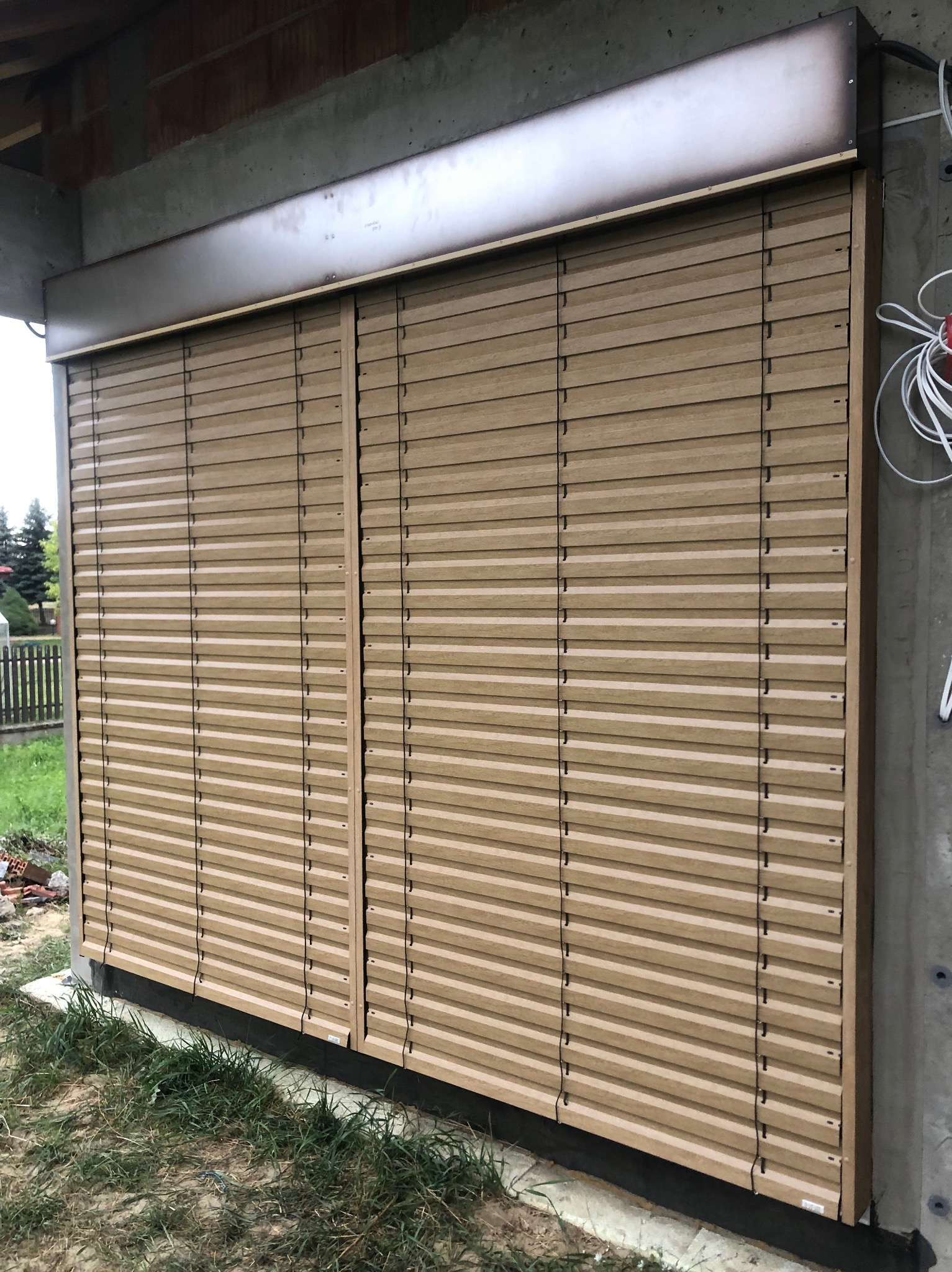 Zamknięte żaluzje fasadowe w kolorze drewna, zamontowane na zewnątrz budynku w trakcie budowy. Widoczne przewody elektryczne i elementy konstrukcyjne.