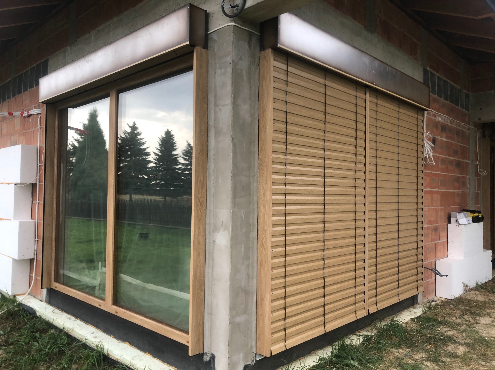 Drewniane żaluzje fasadowe zamontowane na oknie budynku w trakcie budowy. Widoczne niedokończone ściany z cegły i styropian. Drewniana rama okna i szare elementy konstrukcyjne.