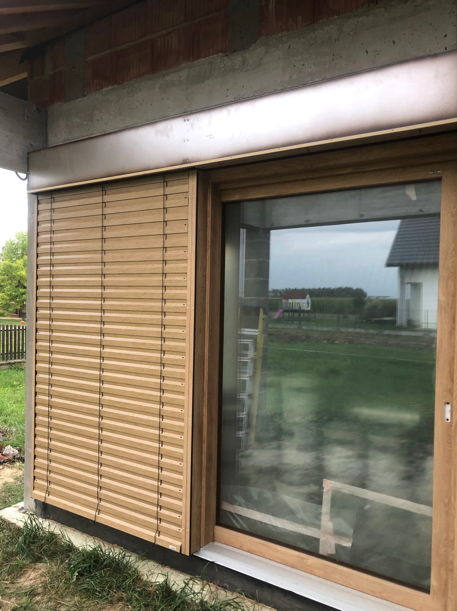 Zamknięta, beżowa roleta zewnętrzna zamontowana obok okna z drewnianą ramą w surowym stanie budynku. W tle widok na zielone pole i dom.