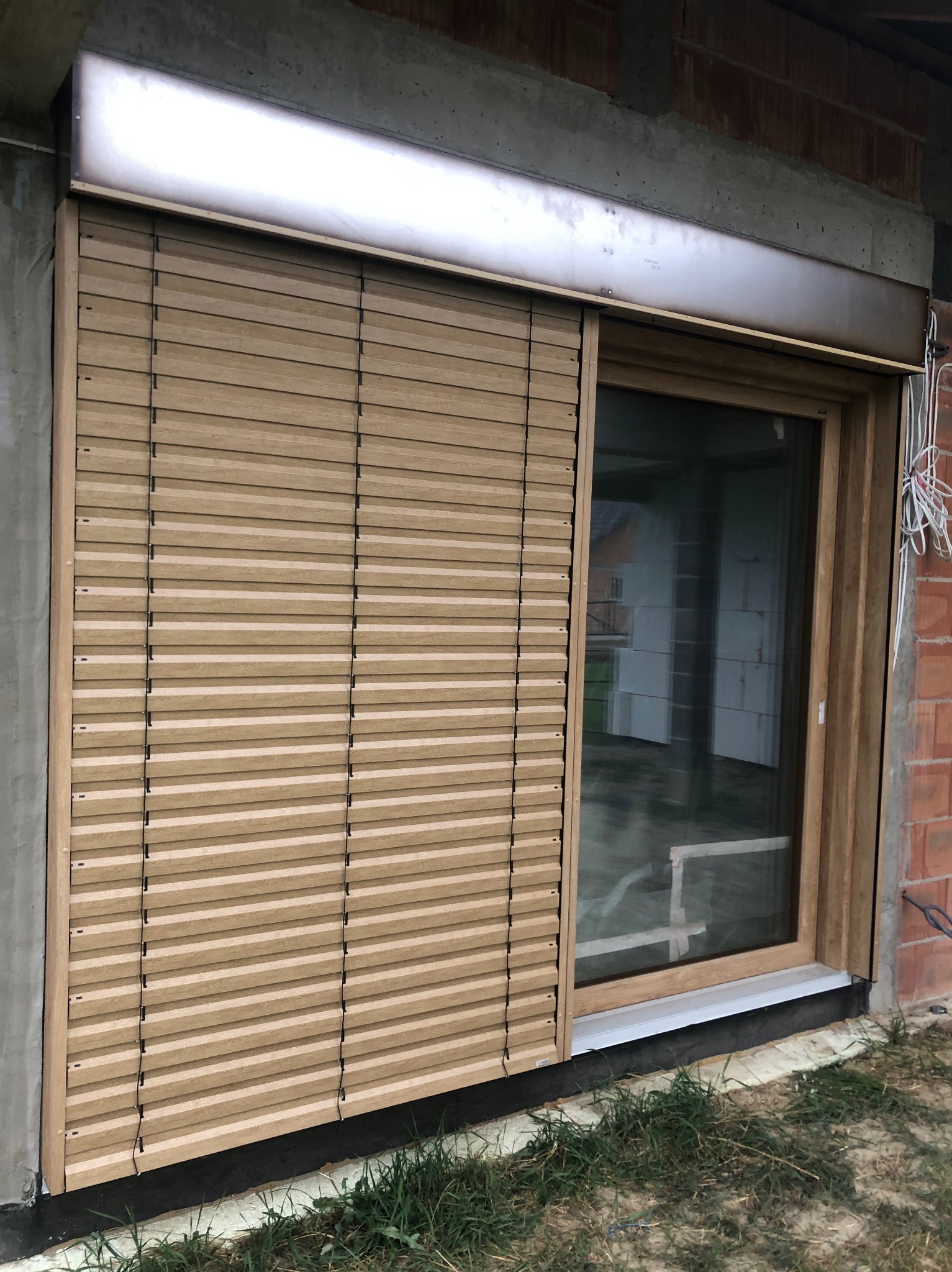 Drewnopodobna roleta zewnętrzna zamontowana na oknie w surowym stanie budynku, z widocznym fragmentem elewacji i trawnikiem. Częściowo zasłonięta szyba okna.
