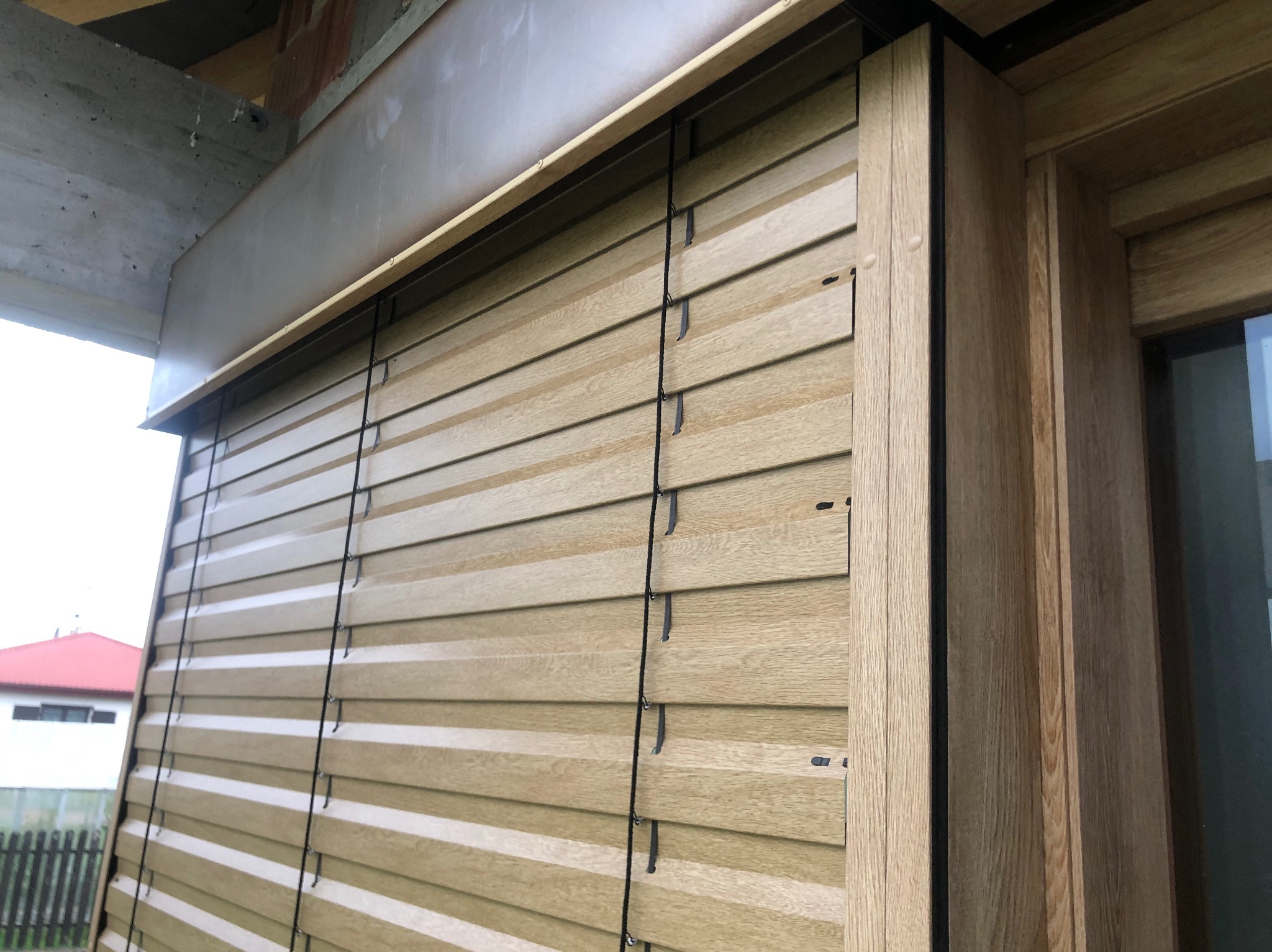 Zamknięte żaluzje drewniane w kolorze jasnego dębu, widoczne sznurki regulacyjne, zarys okna po prawej stronie, fragment budynku w tle, ujęcie z dołu.