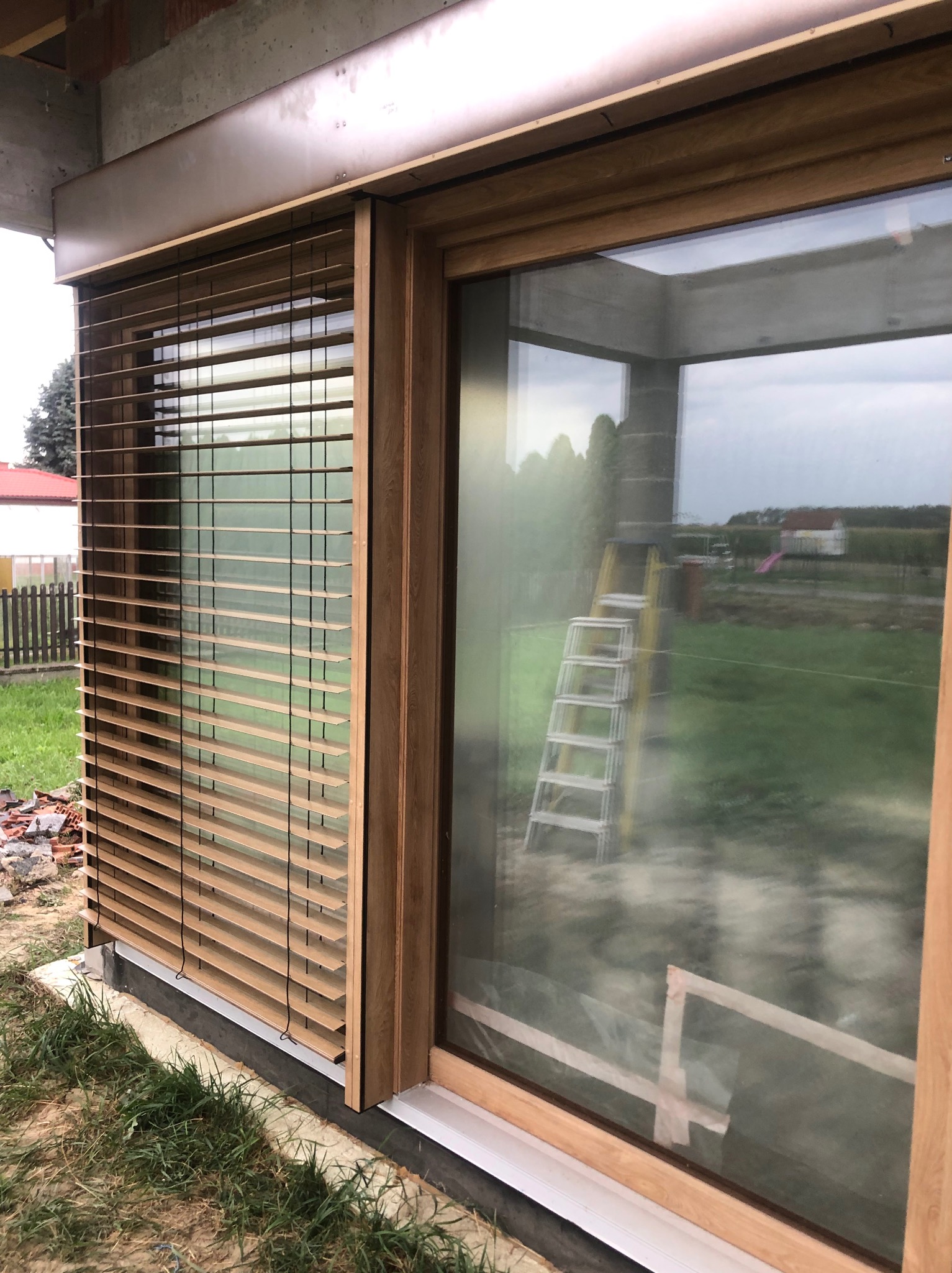 Zamknięte żaluzje drewniane w oknie z drewnianą ramą na tle trawnika i budynków. Widoczna drabina oparta o szybę okna.