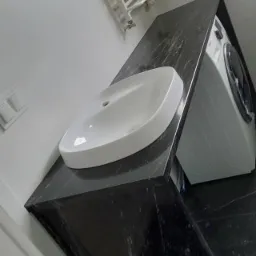 Biała umywalka nablatowa na czarnym, błyszczącym blacie w łazience, obok pralka, widoczny grzejnik drabinkowy i okno.