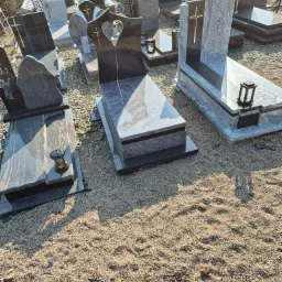 Różnorodne, gotowe pomniki z ciemnego i jasnego granitu, niektóre z wyrytymi krzyżami i lampionami, ustawione na żwirowym placu w zakładzie kamieniarskim.