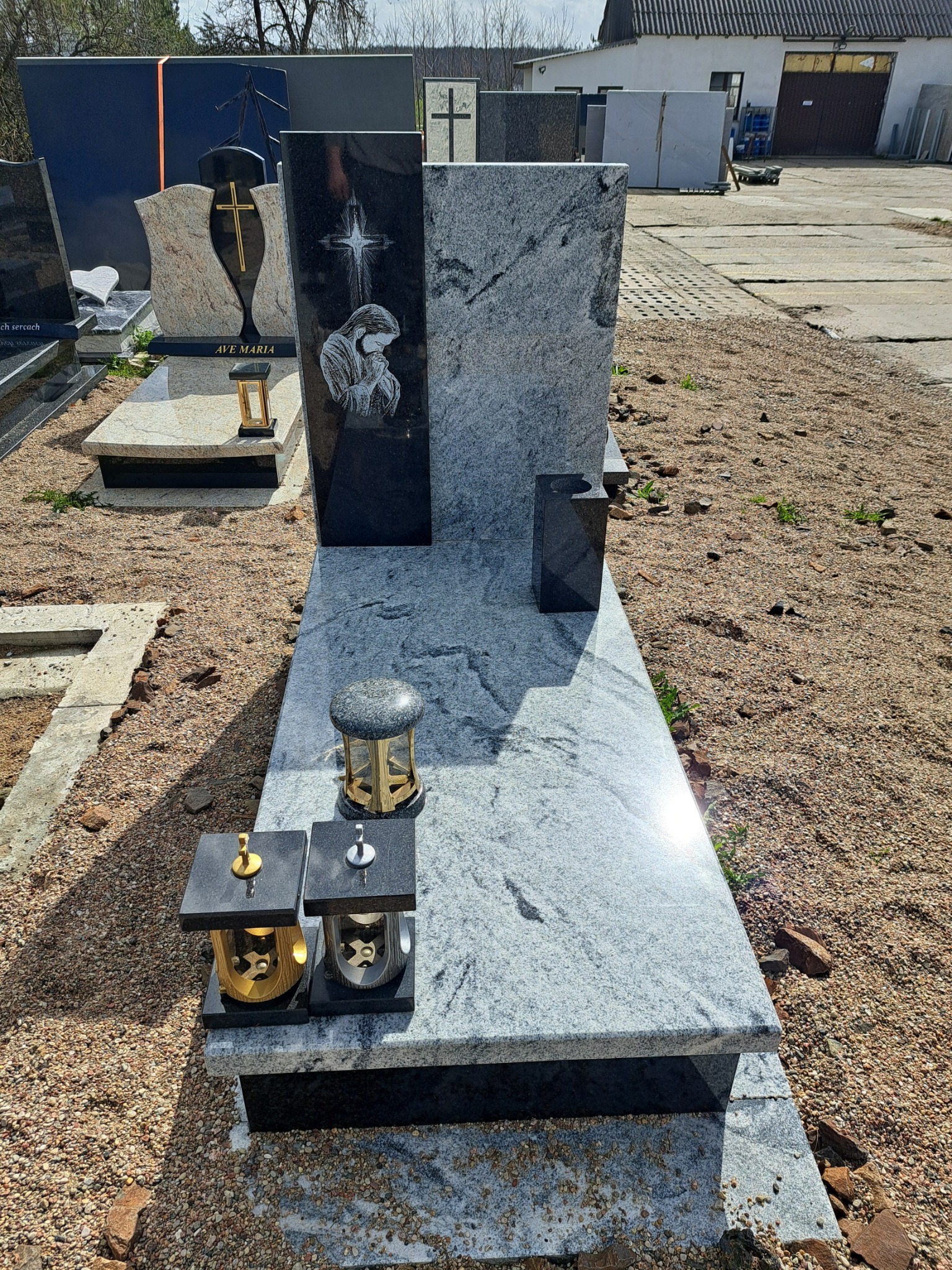 Jasnoszary nagrobek z czarnymi elementami, z wygrawerowaną postacią Matki Boskiej, ozdobiony lampionami, na żwirowym podłożu cmentarza.