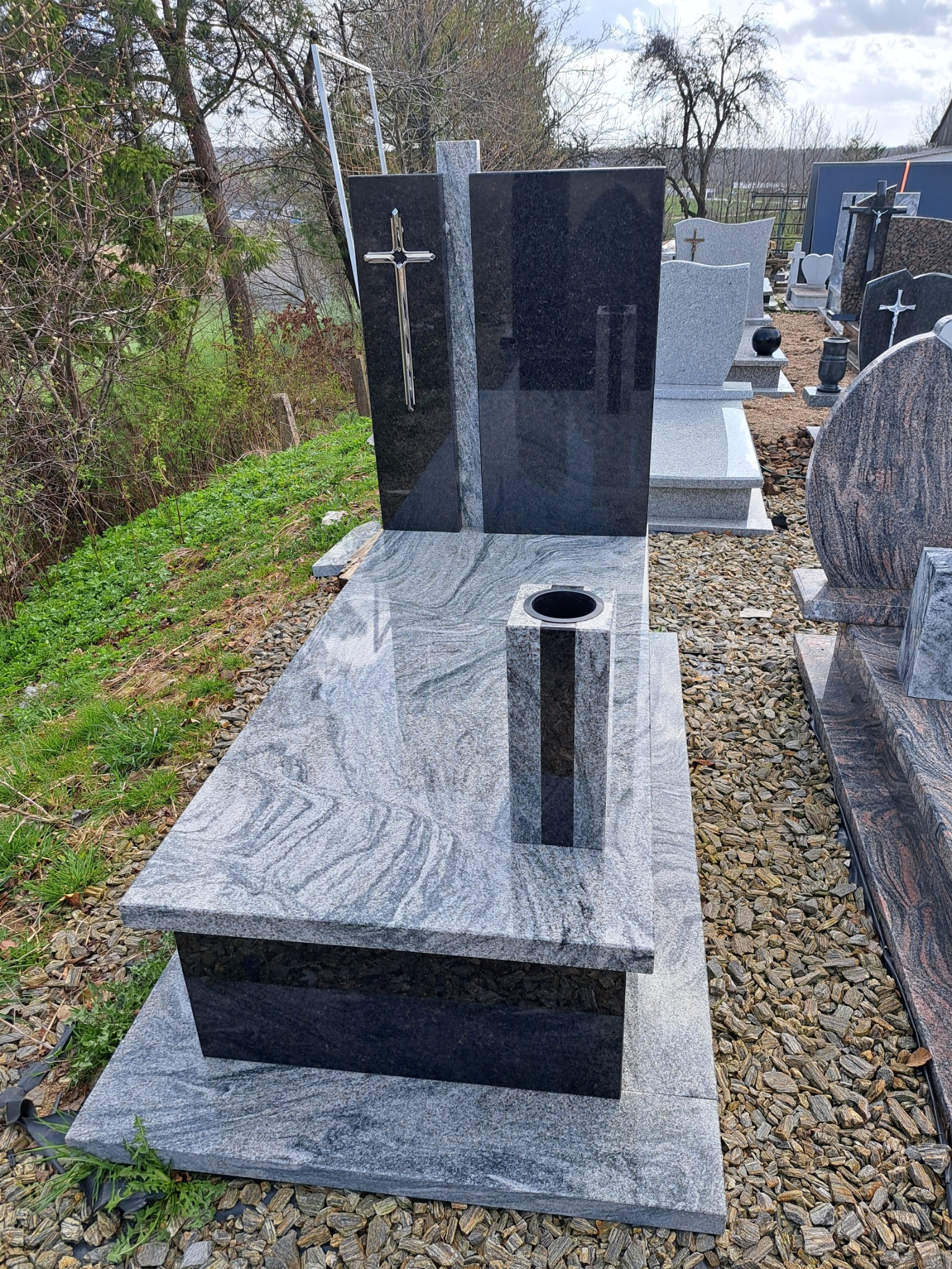 Nowoczesny nagrobek z szarego granitu z czarnymi elementami, w tym wazonem i krzyżem, otoczony żwirem i zielenią.