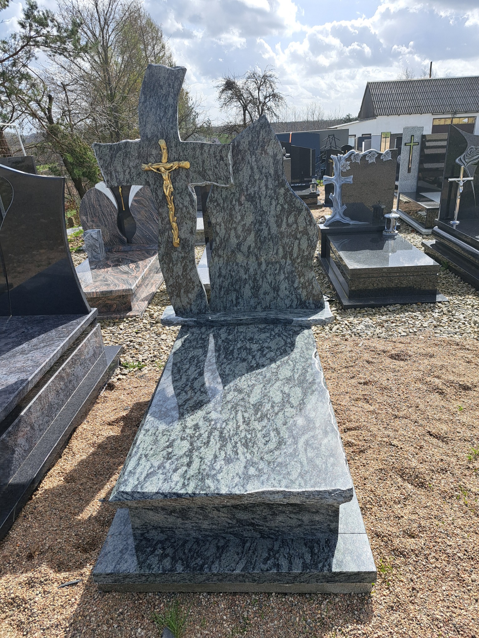 Nowoczesny, dwupoziomowy nagrobek z szarego kamienia z krzyżem i złotym Jezusem, ustawiony na żwirowym podłożu wśród innych nagrobków w słoneczny dzień.