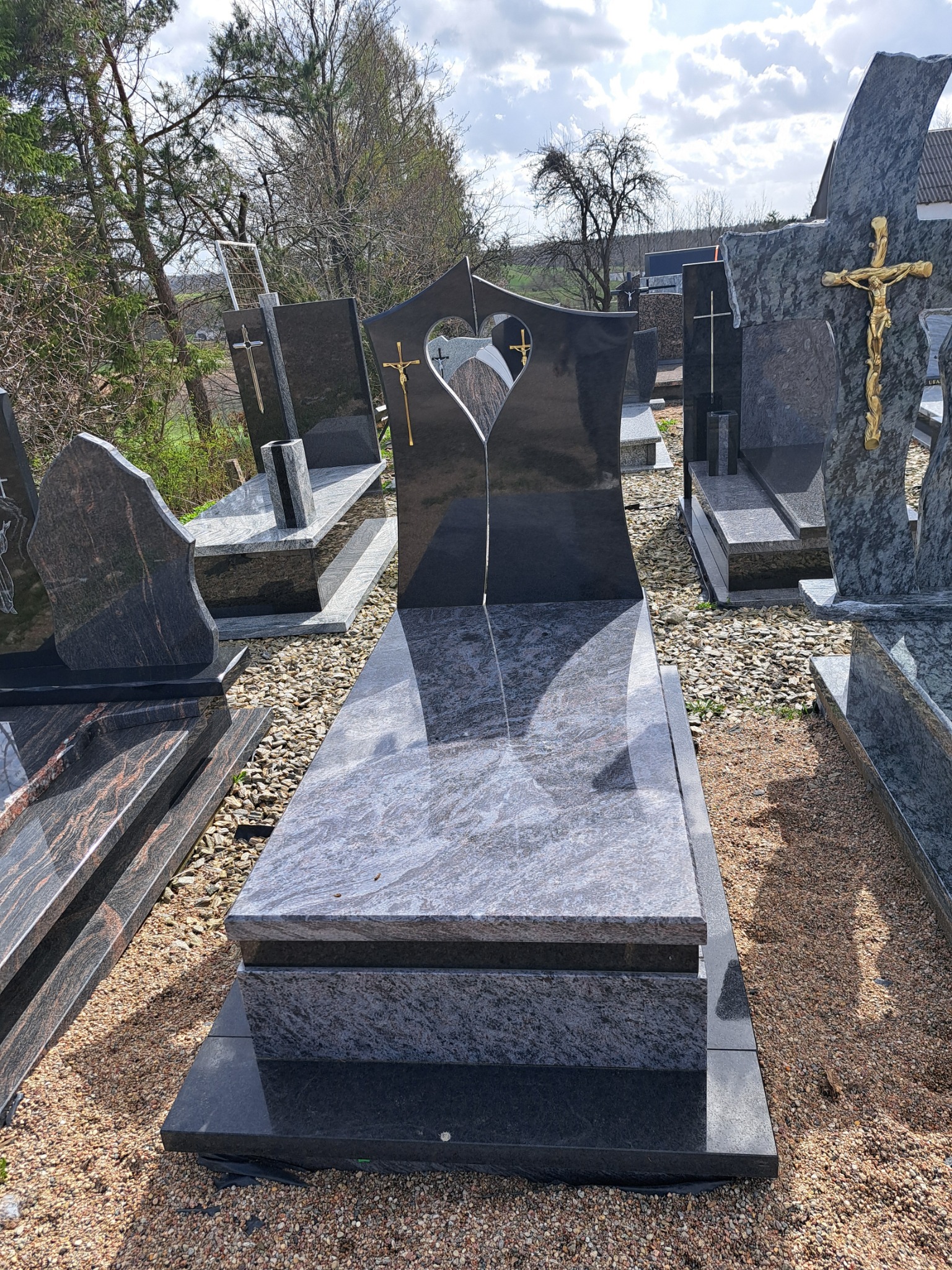 Wystawa nagrobków z ciemnego granitu, jeden z wyciętym sercem i złotym krzyżem, ustawione na żwirowym podłożu w pochmurny dzień.