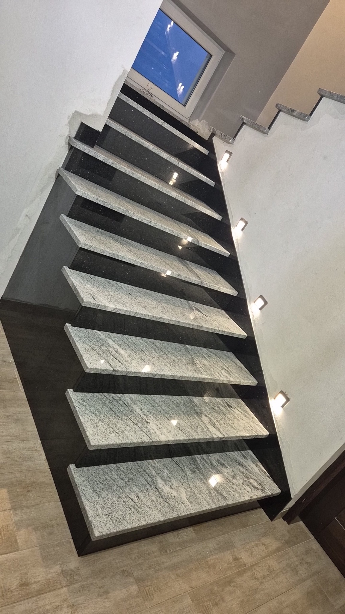 Nowoczesne schody wewnętrzne z szarych stopni kamiennych na tle czarnej ściany, oświetlone małymi punktowymi lampkami, z widokiem na okno dachowe.