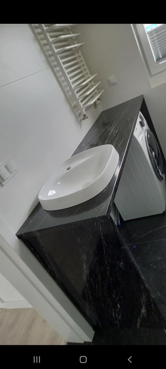 Biała umywalka nablatowa na czarnym, błyszczącym blacie w łazience, obok pralka, widoczny grzejnik drabinkowy i okno.