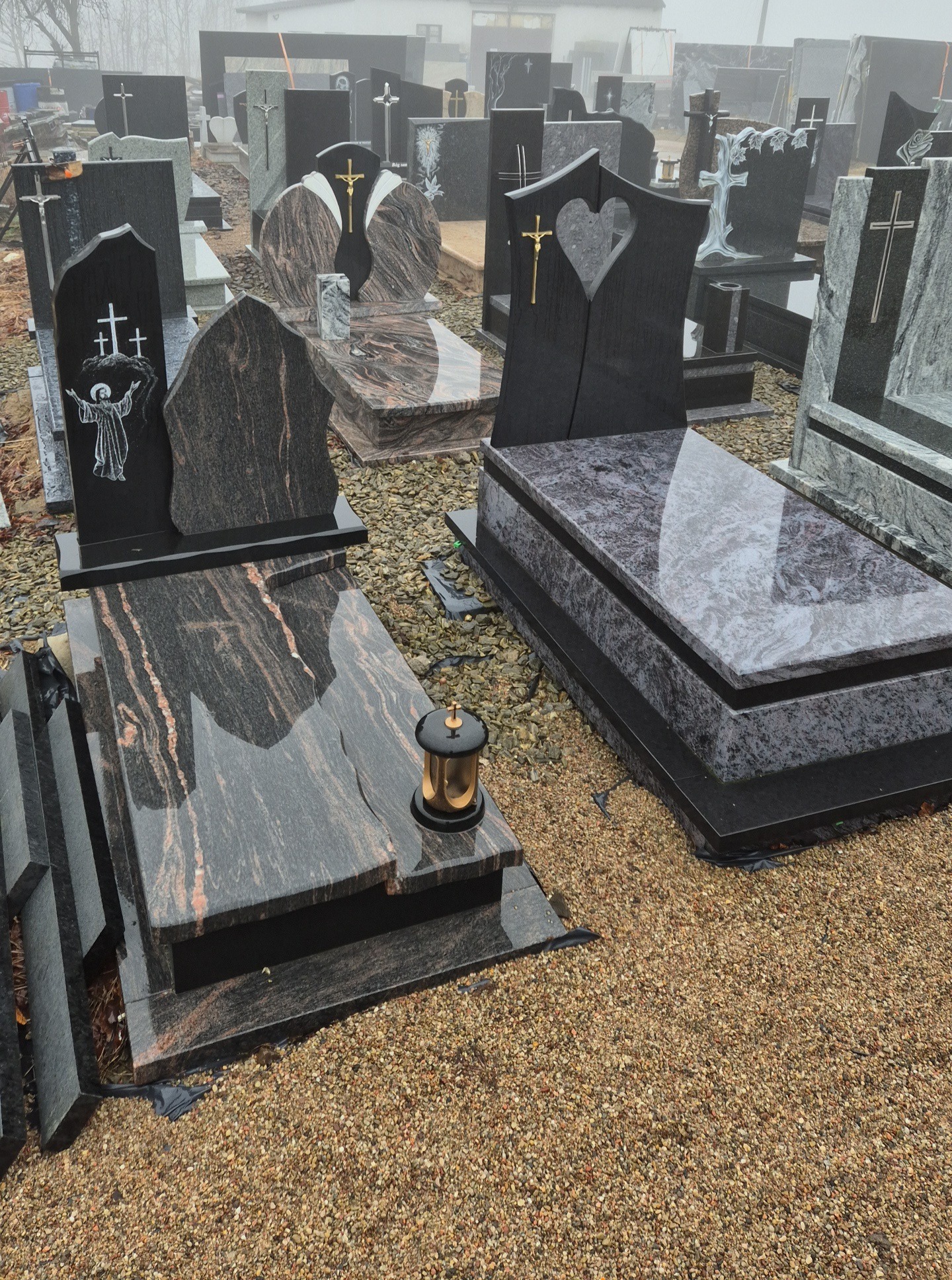 Różnorodne, wypolerowane pomniki z ciemnego kamienia na żwirowym podłożu, niektóre z krzyżami i ornamentami, w tle mgła.
