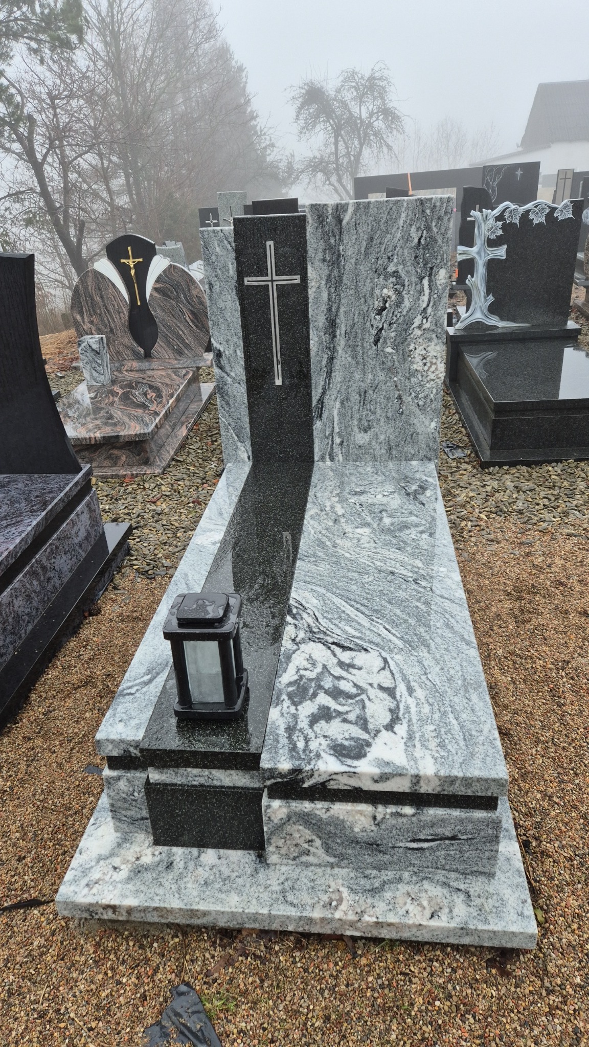 Jasnoszary nagrobek z czarnym elementem centralnym z wyrytą srebrną krzyżem, na cmentarzu w mglisty dzień, z lampionem na płycie.