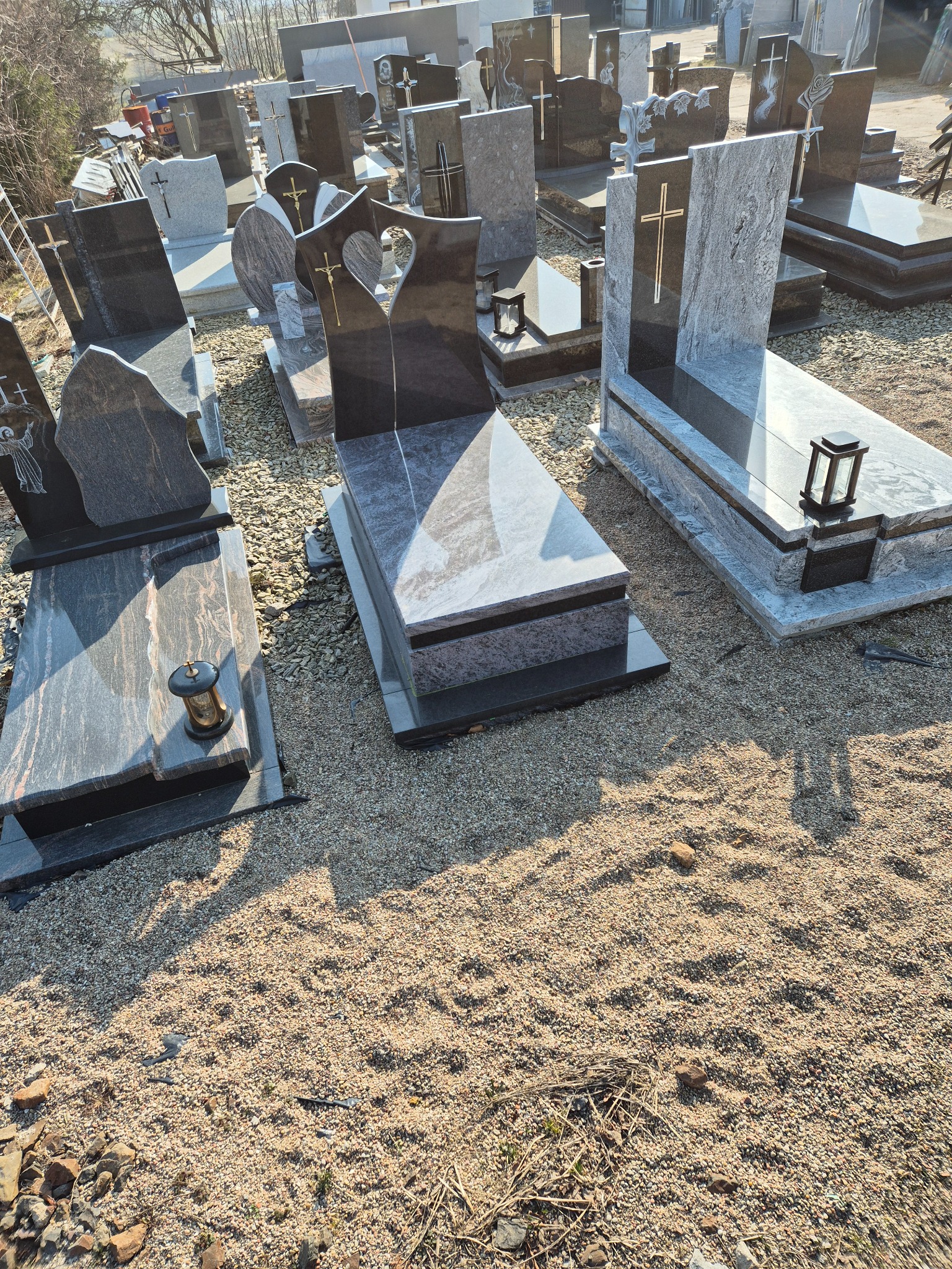 Różnorodne, gotowe pomniki z ciemnego i jasnego granitu, niektóre z wyrytymi krzyżami i lampionami, ustawione na żwirowym placu w zakładzie kamieniarskim.