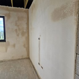Zakład Ogólnobudowlany Jarosław Witek - Wnętrze surowego pomieszczenia po nałożeniu tynków maszynowych, z widocznymi niedoskonałościami i nierównomiernym kolorem, przygotowane do dalszych prac wykończeniowych, z widoczną instalacją...