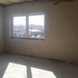 Pokój po nałożeniu tynków maszynowych, widoczne dwa okna z białymi ramami i surowy strop z widocznymi niedoskonałościami.