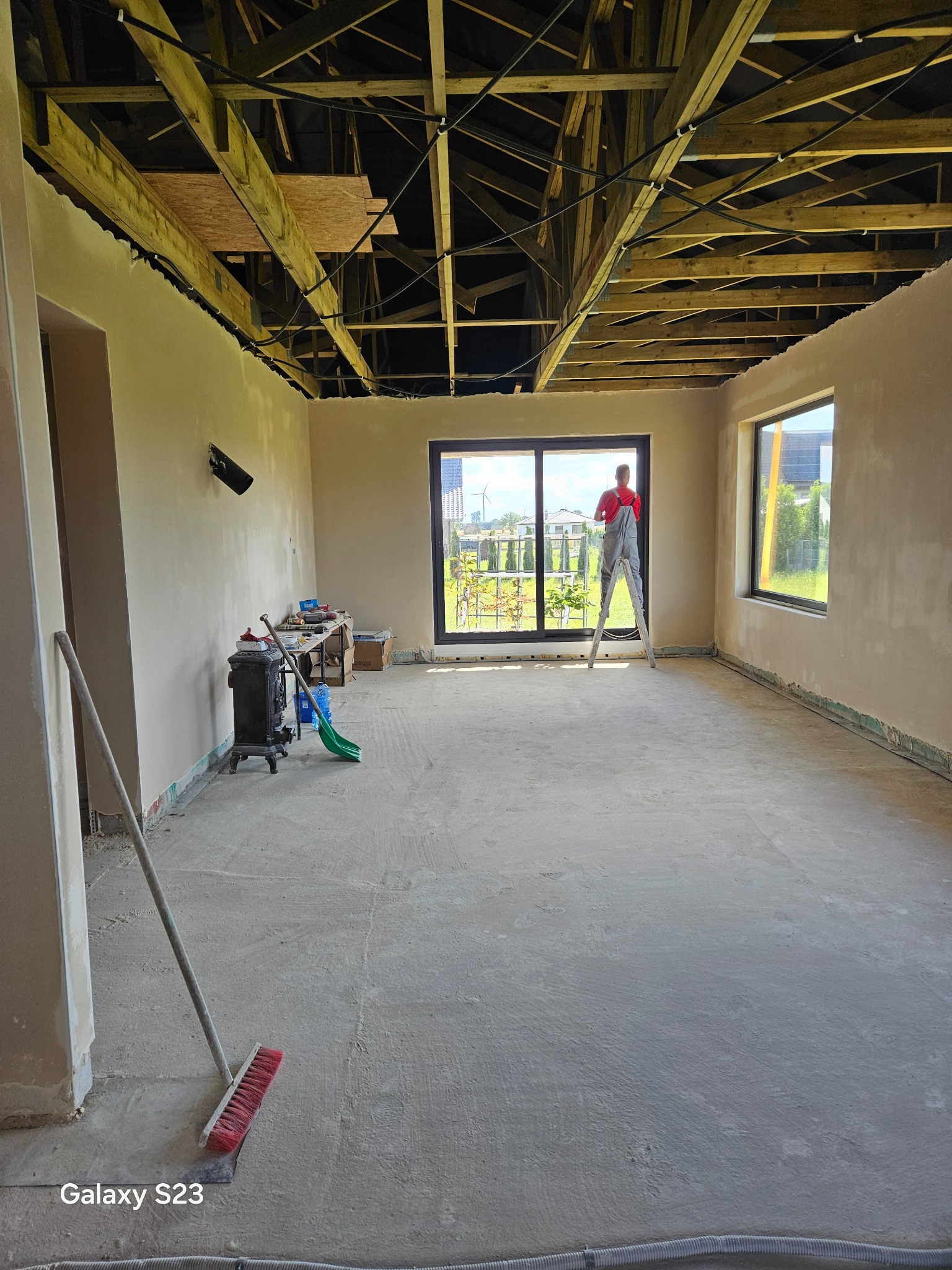 Wnętrze w trakcie prac wykończeniowych: ściany po nałożeniu wstępnej warstwy, odsłonięta konstrukcja drewniana sufitu, pracownik na drabinie przy oknie, widoczne narzędzia i akcesoria malarskie.