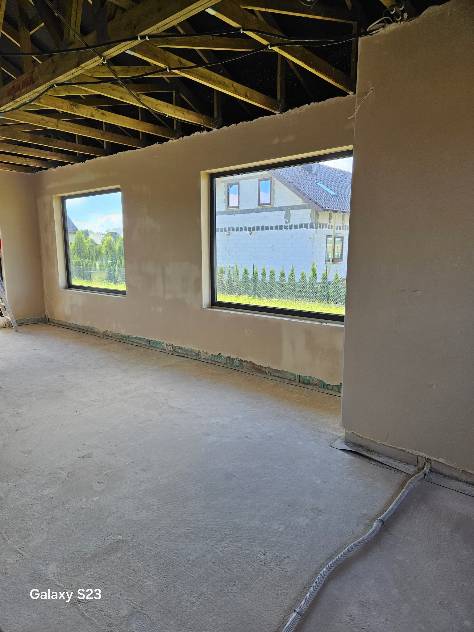 Wnętrze w trakcie prac w Charzynie: ściany pokryte świeżym tynkiem, widoczne dwa okna z widokiem na dom i zieleń, surowy sufit z drewnianą konstrukcją, szary beton na podłodze, rury instalacyjne...