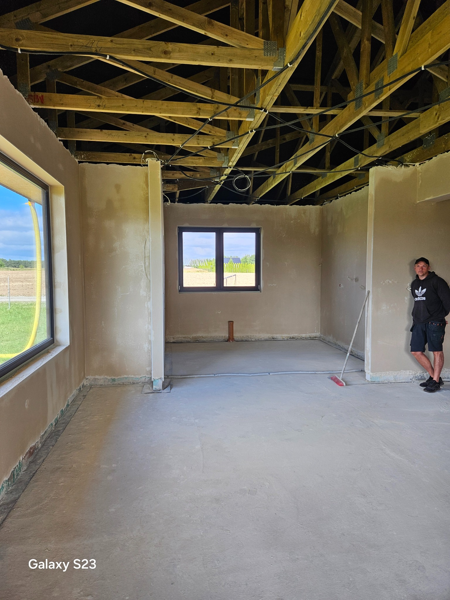 Wnętrze domu w stanie deweloperskim po nałożeniu tynków, widoczny pracownik oparty o ścianę, surowa drewniana konstrukcja dachu, okna z widokiem na zewnątrz, podłoga przygotowana do dalszych prac...