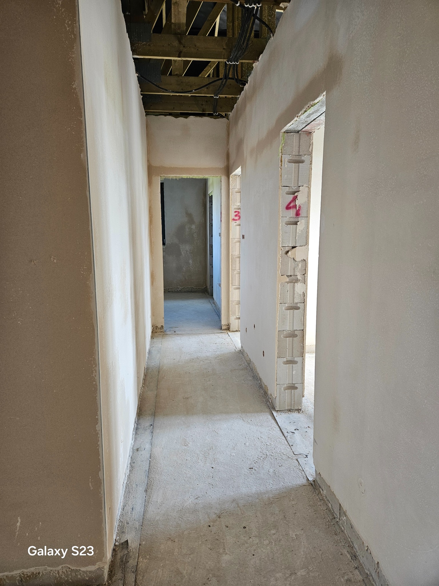 Wnętrze budynku w trakcie prac wykończeniowych: ściany pokryte świeżym, jasnym tynkiem, widoczne surowe belki stropowe i instalacje, ślady czerwonych oznaczeń na bloczkach konstrukcyjnych w przejściu.