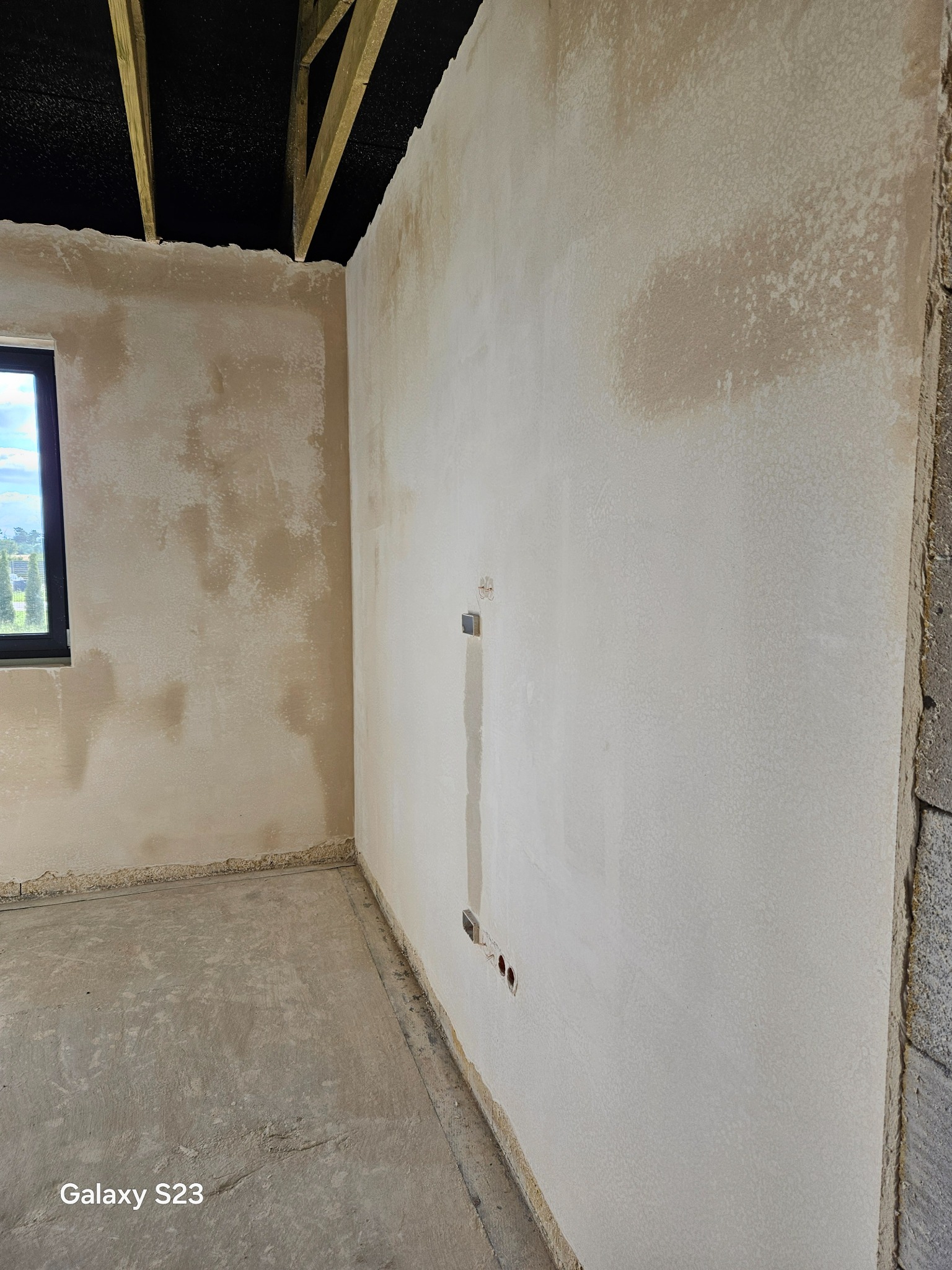 Wnętrze surowego pomieszczenia po nałożeniu tynków maszynowych, z widocznymi niedoskonałościami i nierównomiernym kolorem, przygotowane do dalszych prac wykończeniowych, z widoczną instalacją...