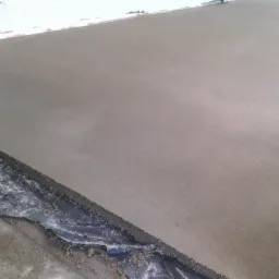 Świeżo wylana, gładka, szara posadzka z widoczną folią ochronną przy ścianie i rurami grzewczymi w tle.