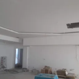 Wnętrze biura po świeżym pomalowaniu na biało, widoczne materiały budowlane i puszki farby w tle, sufit z dekoracyjnym obramowaniem i otworem inspekcyjnym.