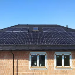 Niedokończony budynek z zamontowanymi panelami słonecznymi na dachu z czarnej dachówki, widoczne trzy okna z folią ochronną.