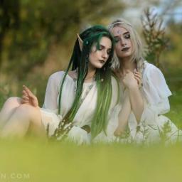 Fotografia Beata Gan - Dwie kobiety w białych sukienkach siedzą na trawie, jedna z zielonymi dredami i spiczastymi uszami, druga z blond włosami i księżycem na czole, obie z makijażem fantasy.