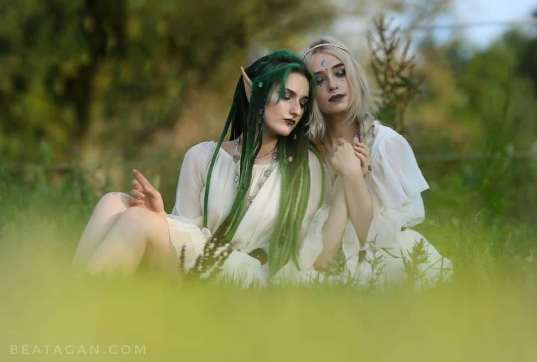 Dwie kobiety w białych sukienkach siedzą na trawie, jedna z zielonymi dredami i spiczastymi uszami, druga z blond włosami i księżycem na czole, obie z makijażem fantasy.