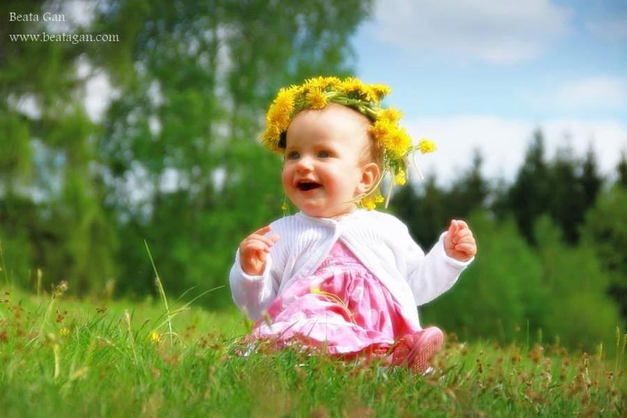 Uśmiechnięta dziewczynka w wianku z żółtych kwiatów siedzi na trawie w słoneczny dzień.