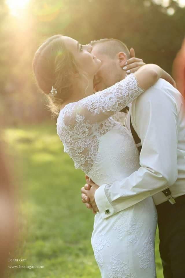 Panna młoda w koronkowej sukni i pan młody obejmują się w kontraście z promieniami słońca w tle.