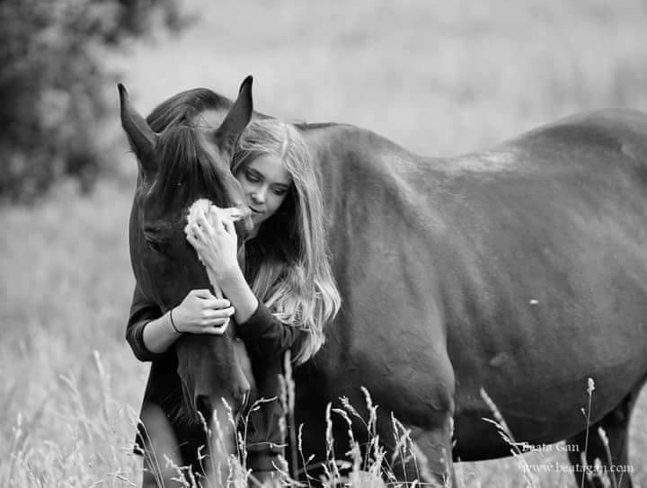 Czarno-białe zdjęcie: młoda kobieta z długimi włosami obejmuje szyję ciemnego konia na łące, wyraz czułości.