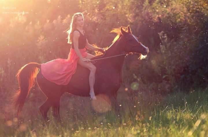 Młoda kobieta w czarnym topie i powiewnej, koralowej spódnicy, boso jeździ na brązowym koniu przez łąkę w promieniach zachodzącego słońca.