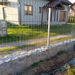 Metalowe ogrodzenie panelowe na niskim murku z kamienia polnego, widok na dom jednorodzinny w słoneczny dzień.