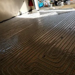 Układanie rur ogrzewania podłogowego na folii z siatką w pomieszczeniu w trakcie budowy, widoczne białe rury w pętlach i elementy instalacji.