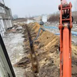 Wąski wykop w ziemi, widoczny z kabiny pomarańczowej koparki, z rurami na dnie wykopu, teren pokryty resztkami śniegu, w tle budynek.