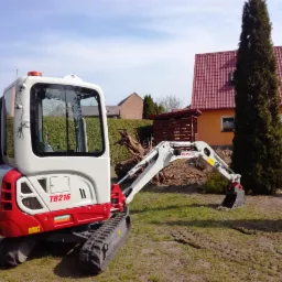 Kompaktowa mini koparka Takeuchi TB216 na trawniku przed domem z pomarańczową elewacją i czerwoną dachówką, widoczne ślady gąsienic na trawie.