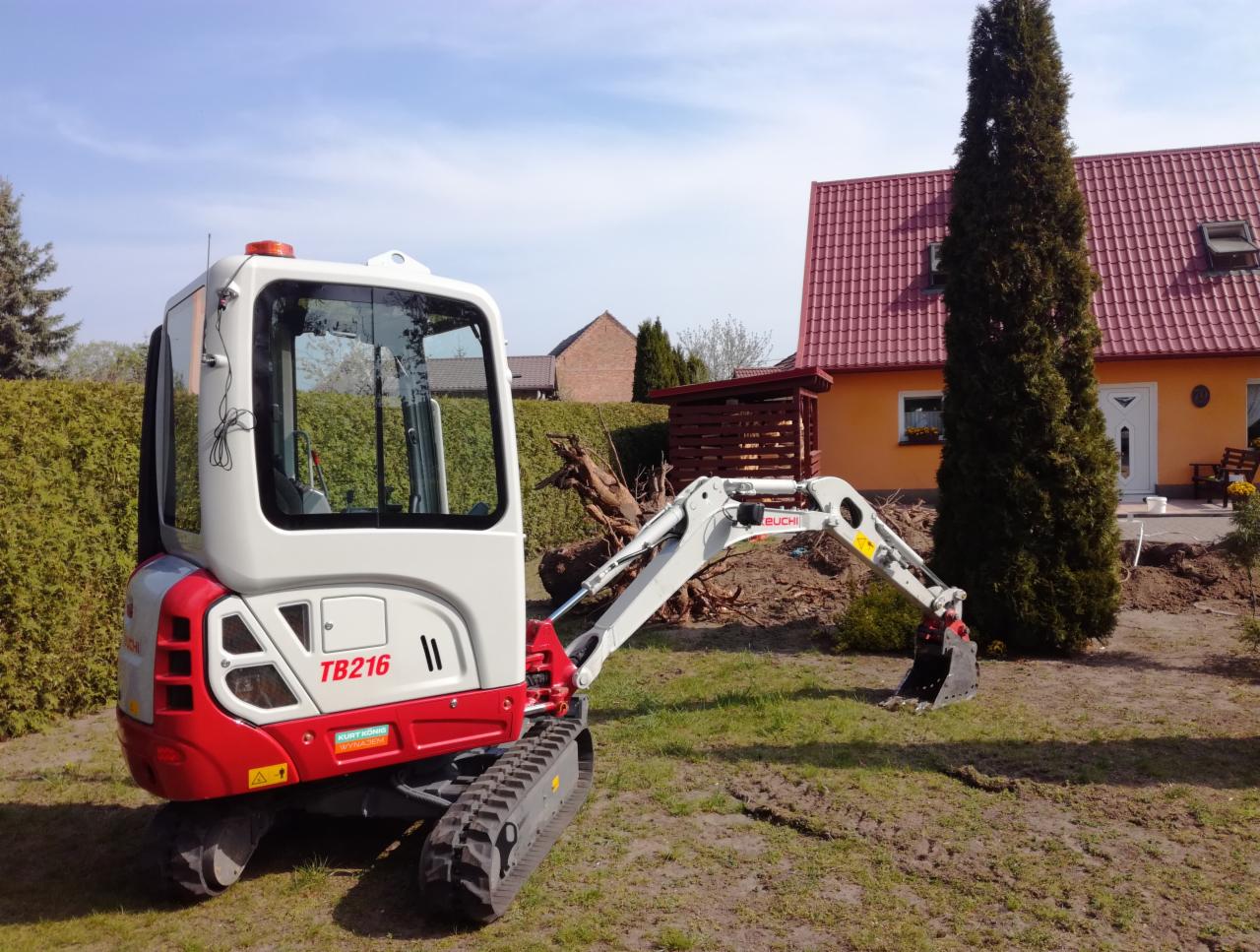 Kompaktowa mini koparka Takeuchi TB216 na trawniku przed domem z pomarańczową elewacją i czerwoną dachówką, widoczne ślady gąsienic na trawie.