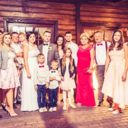 Grupa gości weselnych, w tym para młoda, pozuje do zdjęcia na tle drewnianej ściany z oknem i dekoracjami świetlnymi.