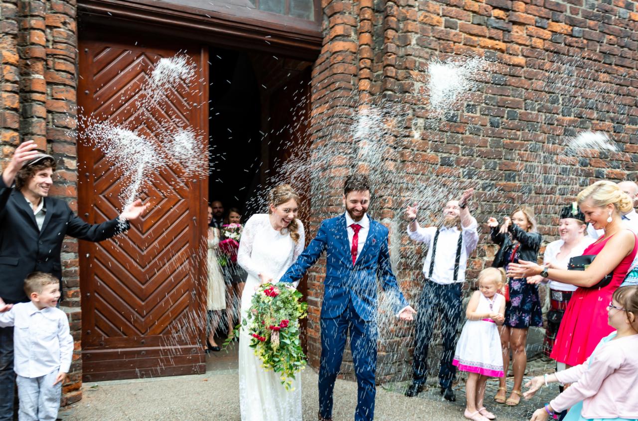 Nowożeńcy w błękitnym garniturze i białej sukni wychodzą z kościoła obsypywani ryżem przez gości weselnych na tle ceglanej ściany i drewnianych drzwi.
