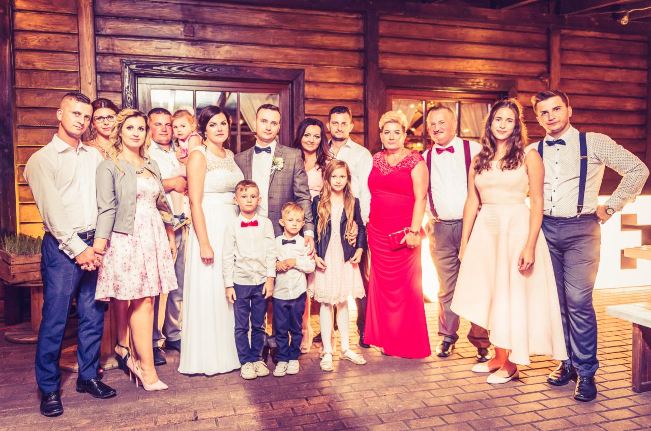 Grupa gości weselnych, w tym para młoda, pozuje do zdjęcia na tle drewnianej ściany z oknem i dekoracjami świetlnymi.